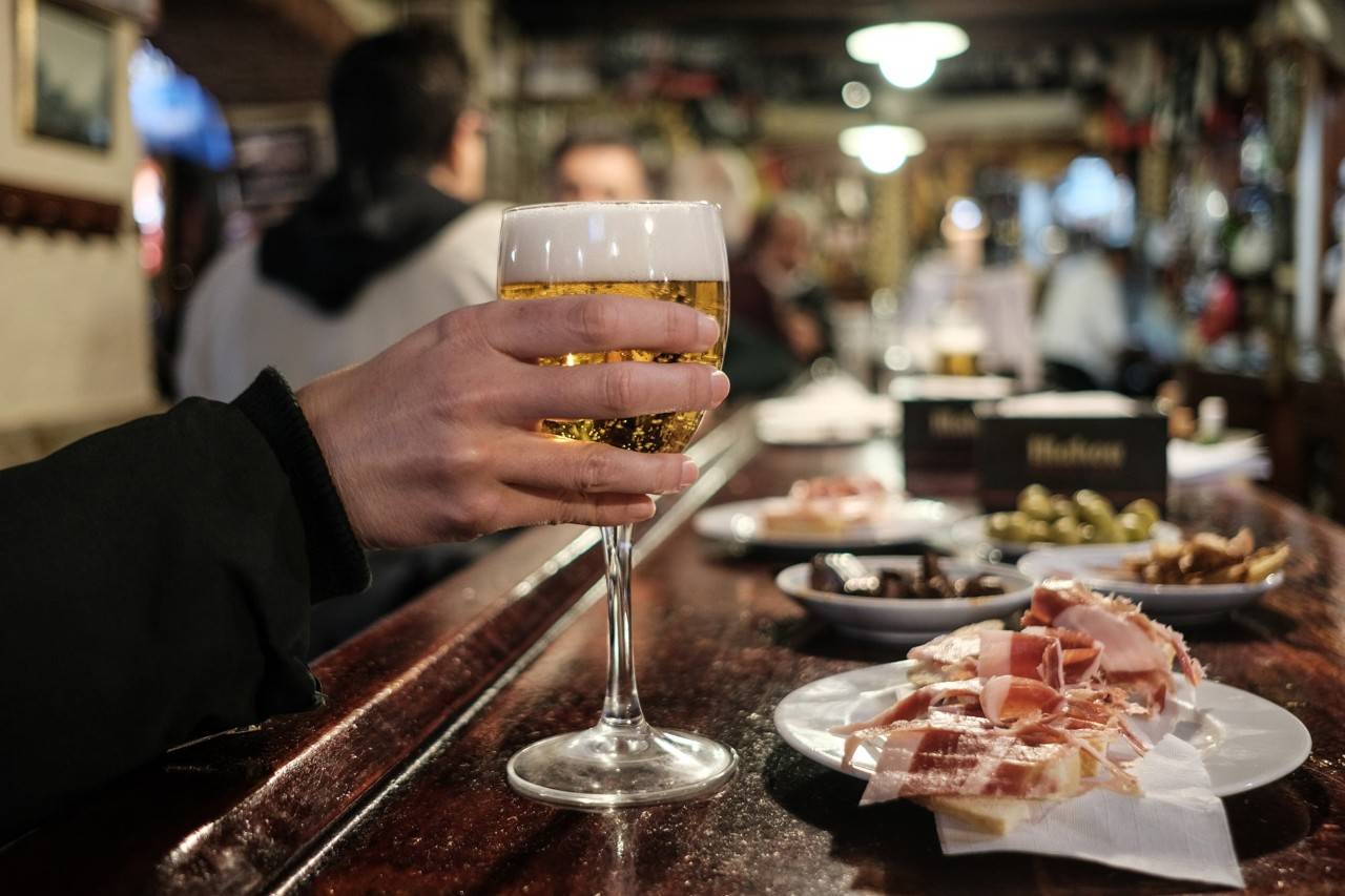 Una mano de mujer sujeta una caña junto a un plato de jamón de la zona en la barra de La Corchuela.