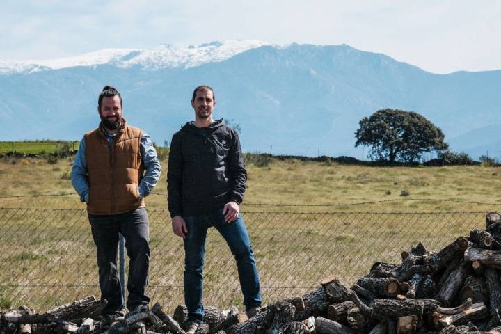 Alfonso y Marcos posan sobre la leña que usarán para destilar su ginebra con las montañas al fondo.