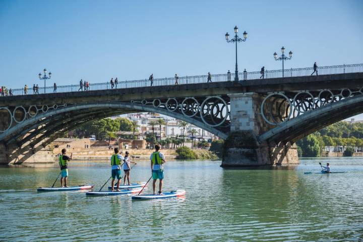 Transeúntes pasean por el icónico Puente de Triana mientras otros pasan por debajo navegando.